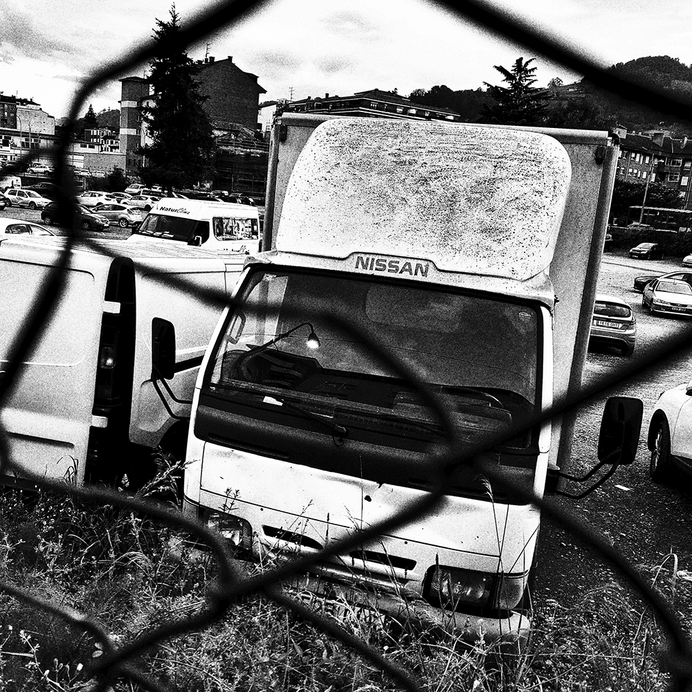 Fotografía urbana en blanco y negro de un camión visto a través de una valla de alambre desenfocada en primer plano, con grano y alto contraste / Urban black and white photograph of a truck seen through a blurred chain-link fence in the foreground, with grain and high contrast. Proyecto / Project: TRASH - Ben Rawman