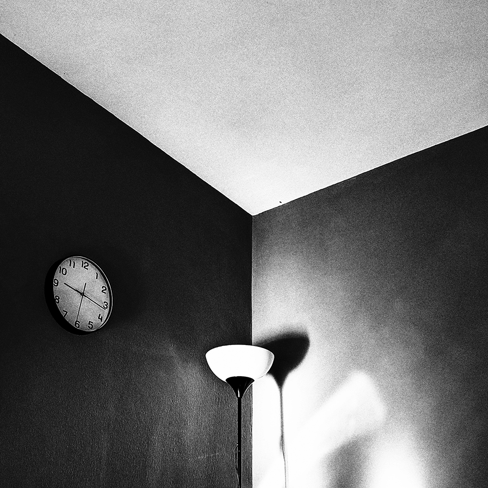 Composición geométrica en blanco y negro de un rincón con reloj de pared, lámpara encendida y fuertes sombras granuladas / Geometric black and white composition of a corner with a wall clock, a lit lamp, and heavy, grainy shadows. Proyecto / Project: TRASH - Ben Rawman