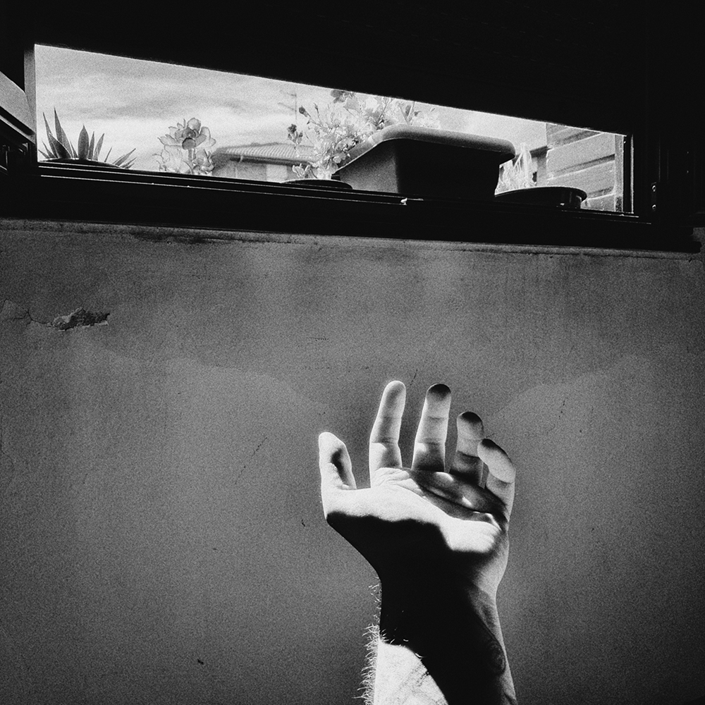 Fotografía expresionista en blanco y negro de una mano alzada hacia una ventana estrecha con plantas, con luces duras y grano dramático / Expressionist black and white photograph of a hand raised toward a narrow window with plants, with harsh lighting and dramatic grain. Proyecto / Project: TRASH - Ben Rawman